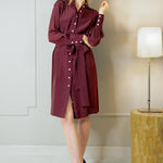 A model wearing a long sleeve, knee-length bordeaux shirtdress with mother-of-pearl buttons and a matching sash belt.