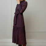 Woman wearing a dark purple print dress with a pattern, standing against a plain wall - by Room 502