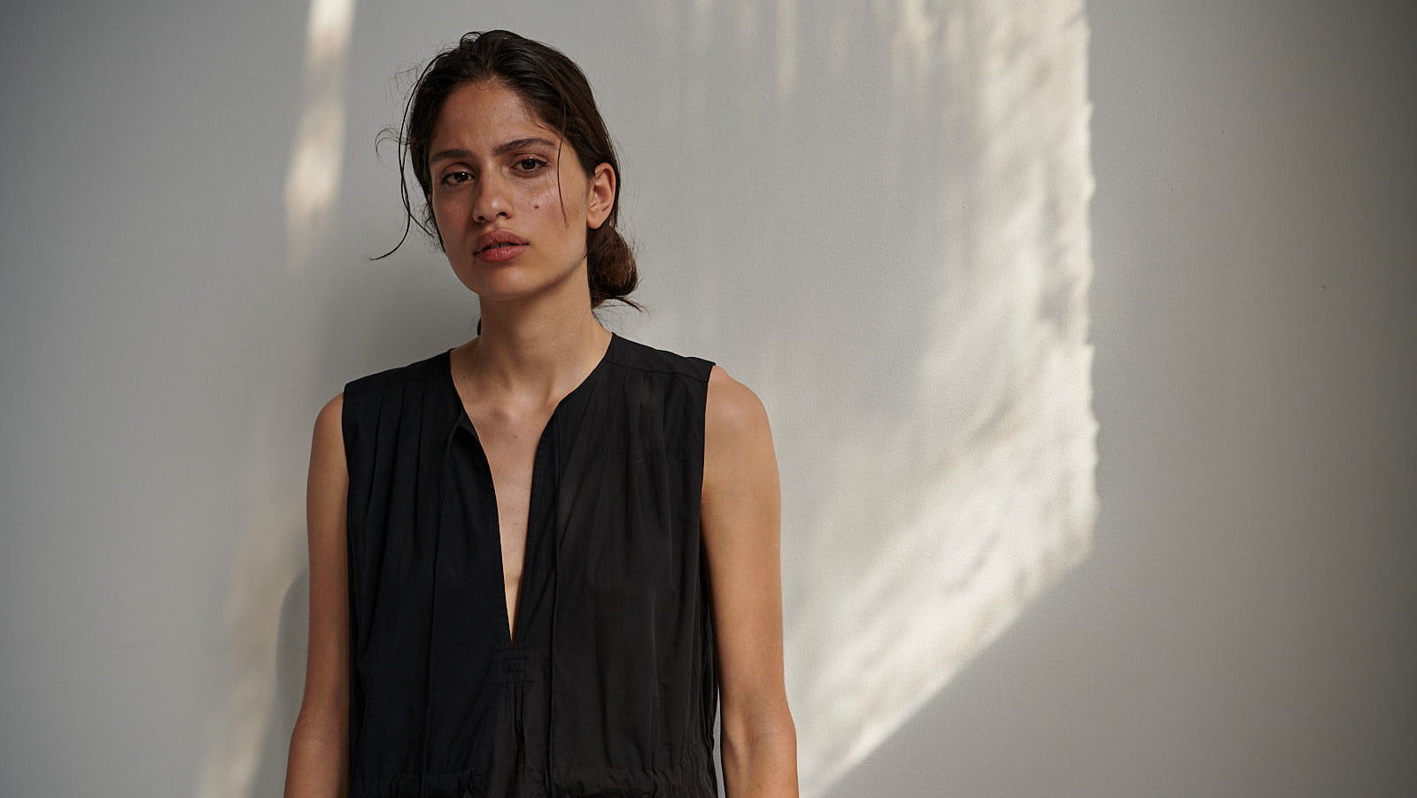 Woman wearing a black sleeveless caftan dress against a light gray wall with shadows.