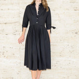 Woman wearing a black shirtdress against a light wooden paneled background